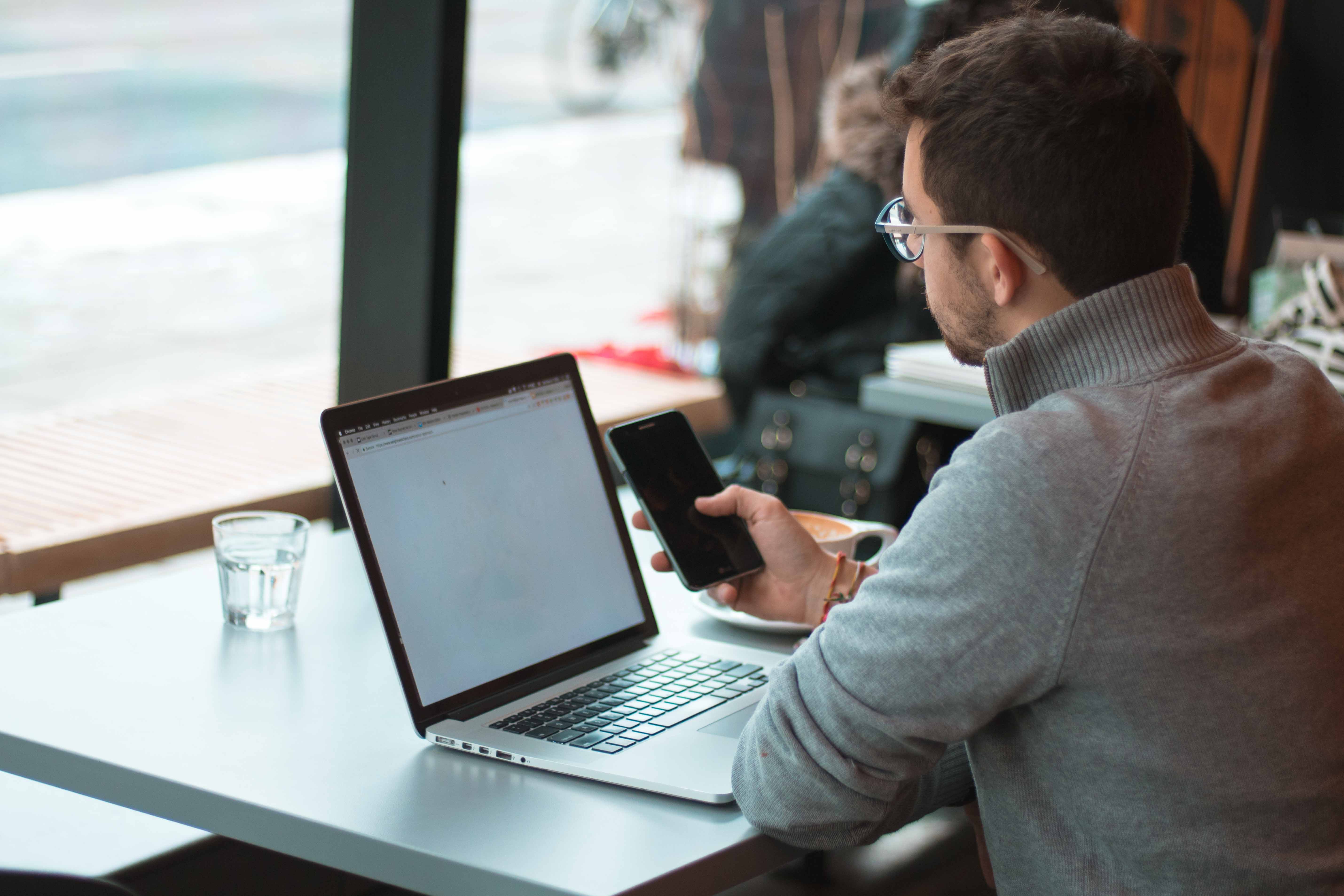 Man sat down at a coffee shop looking through his phone and laptop